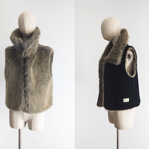 May include: Two vests on mannequins. One is a faux fur vest in shades of brown and tan, with a high collar. The other is a black knit vest with a brown faux fur collar and trim. Both vests are sleeveless.