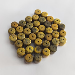 May include: A collection of yellow and dark green cylindrical beads with a hole through the centre. The beads have a swirled pattern and are arranged on a white surface. The beads are a mix of yellow and dark green.