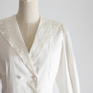 May include: A white blouse with a double-breasted design and long sleeves. The blouse features a V-neckline with a delicate, embroidered lace collar. The buttons are white and round.