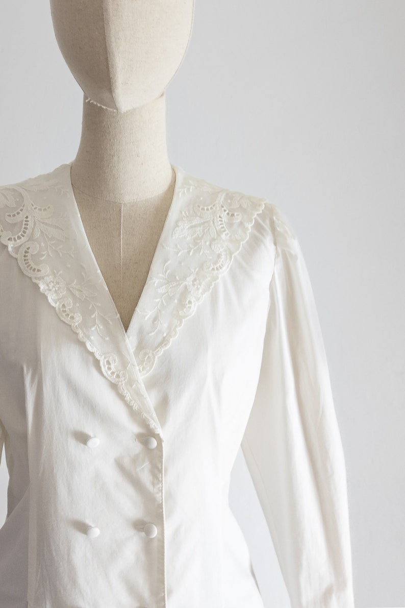 May include: A white blouse with a double-breasted design and long sleeves. The blouse features a V-neckline with a delicate, embroidered lace collar. The buttons are white and round.