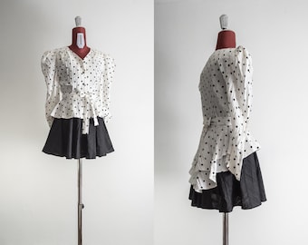 May include: A vintage two-piece outfit on a mannequin. The top is white with black polka dots, a V-neck, and puffed sleeves. It has a waist tie. The skirt is black and flared. The outfit is displayed against a white background.