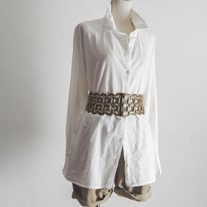 Op de afbeelding: Een wit, lang mouwig overhemd met knopen en een geweven riem. De riem is bruin met gouden accenten.