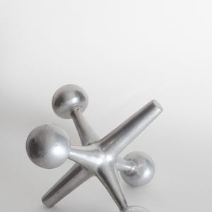 K&ouml;nnte beinhalten: Eine silberfarbene Metallskulptur in Form eines Spiel-Jacks, bestehend aus einem zentralen Knotenpunkt mit sechs strahlenf&ouml;rmigen Armen, die jeweils in einer glatten, kugelf&ouml;rmigen Kugel enden. Das Objekt hat ein mattes Metallfinish.
