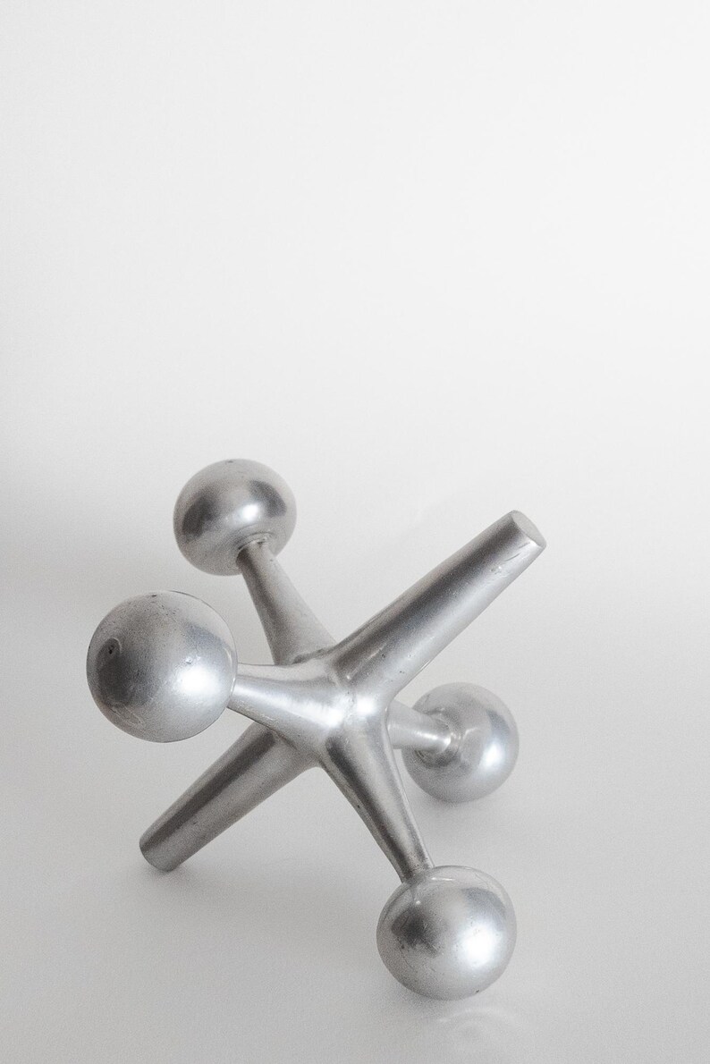 K&ouml;nnte beinhalten: Eine silberfarbene Metallskulptur in Form eines Spiel-Jacks, bestehend aus einem zentralen Knotenpunkt mit sechs strahlenf&ouml;rmigen Armen, die jeweils in einer glatten, kugelf&ouml;rmigen Kugel enden. Das Objekt hat ein mattes Metallfinish.