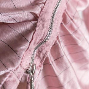 May include: Close-up of a pink fabric with a silver zipper. The fabric has a subtle striped pattern in black. The zipper is partially open, revealing the texture of the material. The overall aesthetic is soft and delicate.