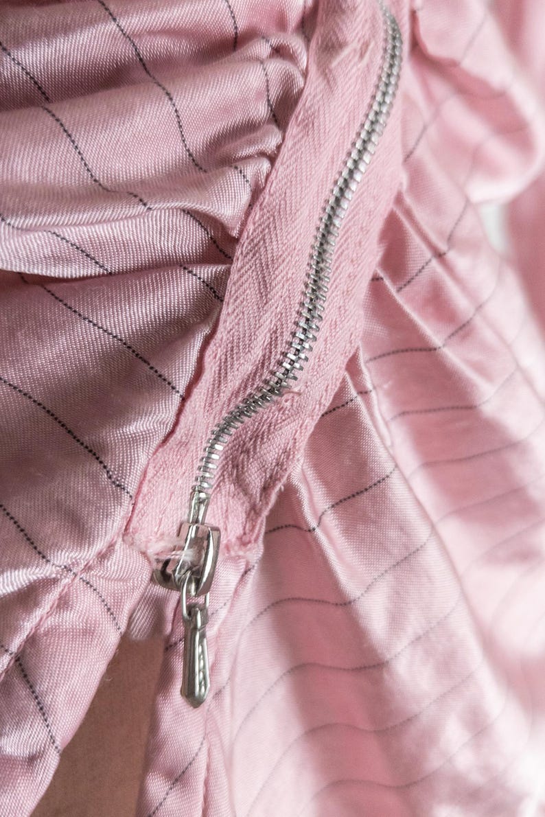May include: Close-up of a pink fabric with a silver zipper. The fabric has a subtle striped pattern in black. The zipper is partially open, revealing the texture of the material. The overall aesthetic is soft and delicate.