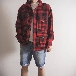 May include: A red and black checked flannel shirt with a button-up front and multiple pockets. The shirt is worn over a white t-shirt and blue denim shorts.