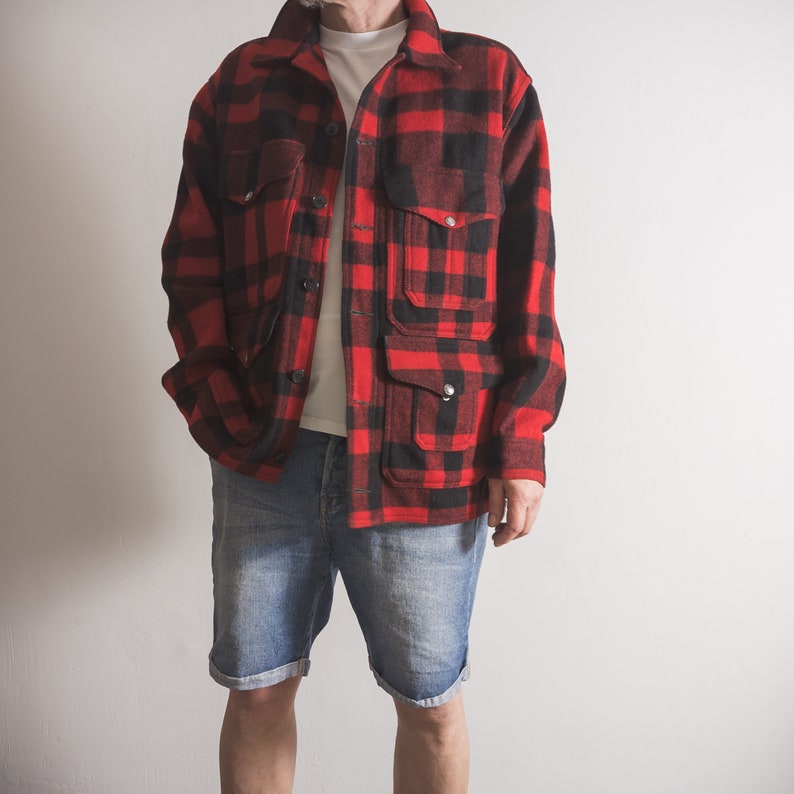 May include: A red and black checked flannel shirt with a button-up front and multiple pockets. The shirt is worn over a white t-shirt and blue denim shorts.