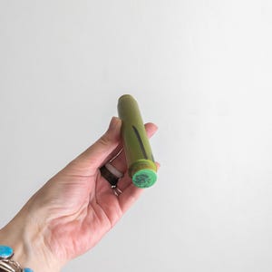 Puede incluir: Un sello cilíndrico verde con una raya negra y una cabeza de sello verde estampada. El sello se sostiene en una mano, con una pulsera turquesa visible en el fondo. El sello probablemente se utiliza para manualidades o proyectos artísticos.