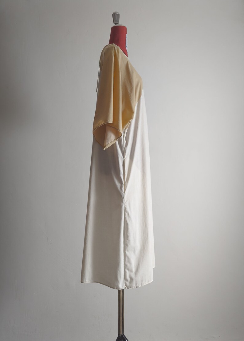 Op de afbeelding: Een witte en beige jurk met een korte mouw op een mannequin. De jurk heeft een losse pasvorm en een eenvoudig ontwerp.