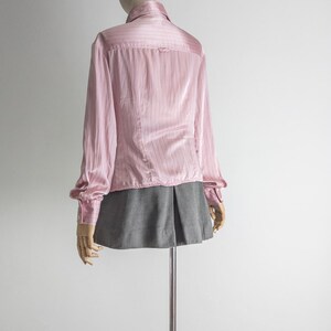 May include: A light pink button-down shirt with vertical stripes and long sleeves. The shirt is paired with a gray pleated skirt. The clothing is displayed on a mannequin against a white background.