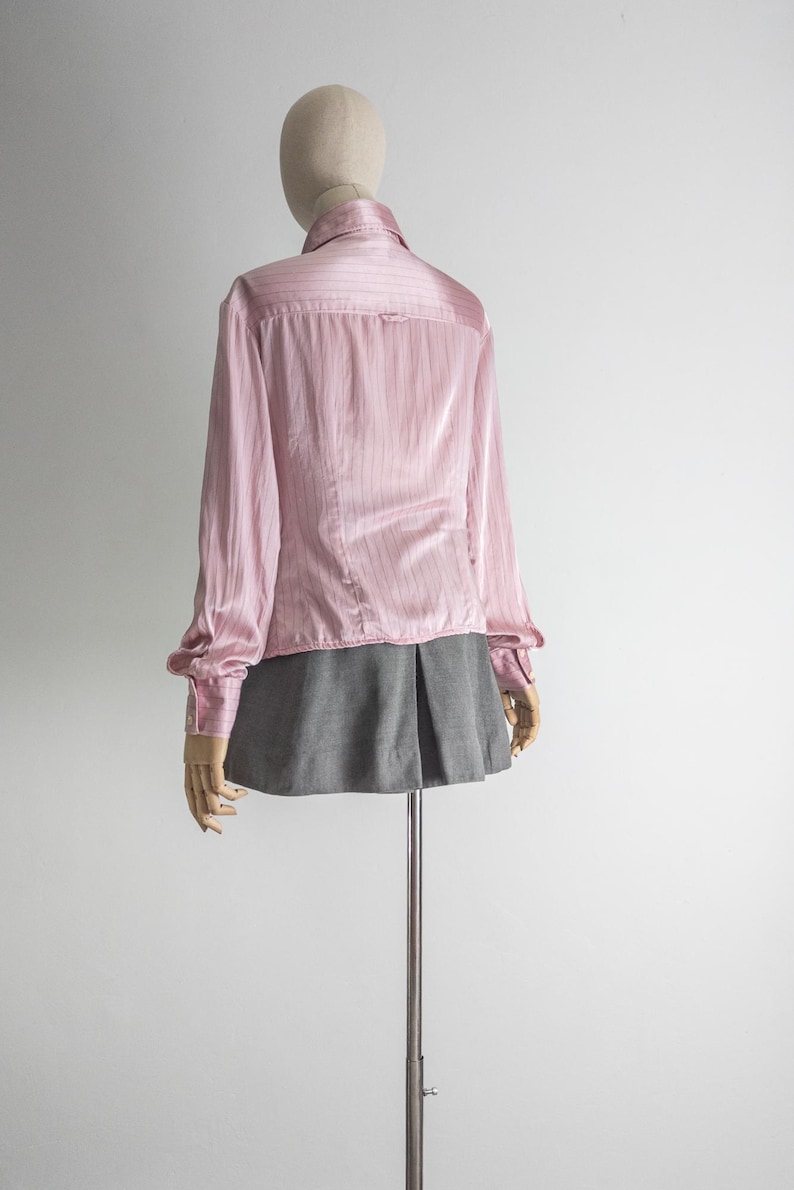 May include: A light pink button-down shirt with vertical stripes and long sleeves. The shirt is paired with a gray pleated skirt. The clothing is displayed on a mannequin against a white background.