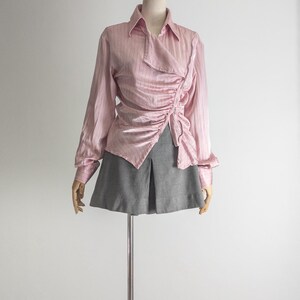 May include: A pink, long-sleeved blouse with a draped, asymmetrical design. The blouse has a collared neckline and is paired with a gray skirt. The outfit is displayed on a mannequin against a white background.