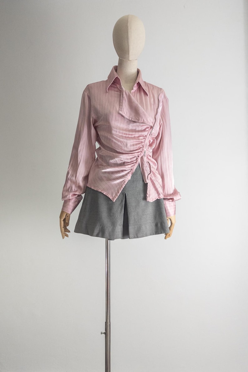 May include: A pink, long-sleeved blouse with a draped, asymmetrical design. The blouse has a collared neckline and is paired with a gray skirt. The outfit is displayed on a mannequin against a white background.