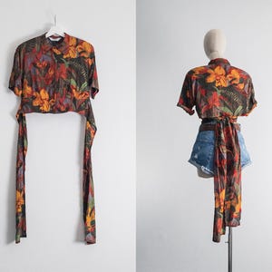 May include: A short-sleeved top with a vibrant floral pattern in shades of orange, red, and green. The top has long, matching fabric ties that hang down. The garment is displayed on a white background.