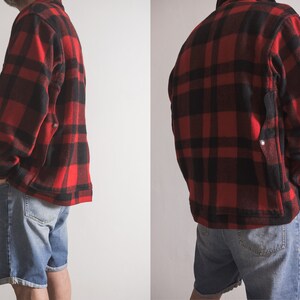 May include: A red and black checked flannel shirt jacket with a patch pocket on the left side. The jacket is worn over a pair of blue denim shorts.
