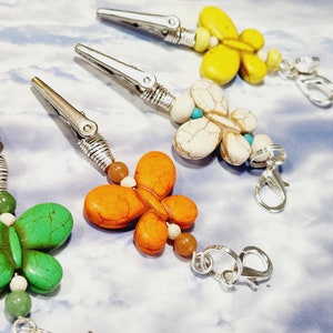May include: Four butterfly-shaped charms with different colored stones, each attached to a silver clip and a silver clasp. The stones are green, orange, white, and yellow.