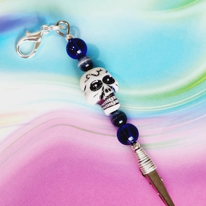 May include: A silver clip with a white skull bead and blue glass beads. The clip is attached to a silver chain with a hook.