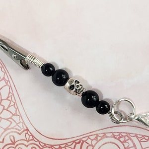 May include: A black and silver beaded clip with a skull charm. The clip is made of metal and has a spring-loaded clasp. The beads are black and round. The skull charm is silver and has a detailed design.