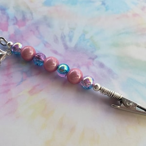 May include: A beaded clip with a silver clasp and a spring-loaded alligator clip. The beads are pink, iridescent, and blue. The clip is designed to hold items.