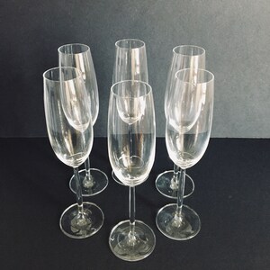 May include: Six clear glass champagne flutes with a tall, slender design. The flutes are arranged in a semi-circle on a dark surface.