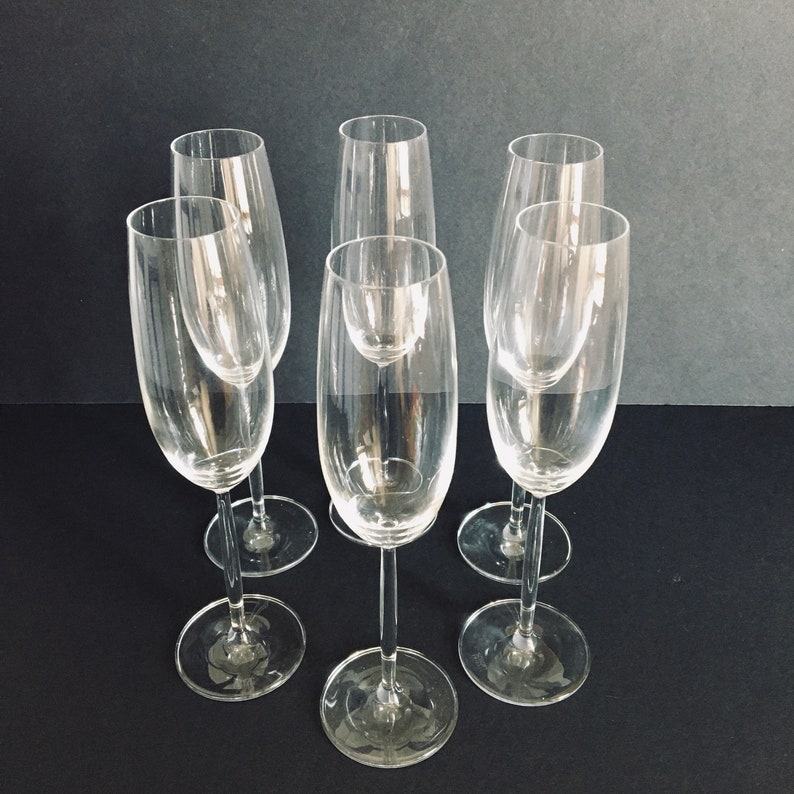 May include: Six clear glass champagne flutes with a tall, slender design. The flutes are arranged in a semi-circle on a dark surface.