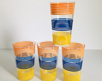May include: Four clear drinking glasses with horizontal bands of orange, blue, and yellow. The glasses are arranged in a pyramid shape, with one glass stacked on top of three others. The glasses are empty and sit on a white surface.
