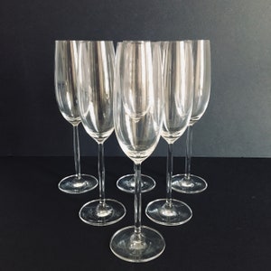 May include: Six clear glass champagne flutes arranged in a semi-circle on a black surface.