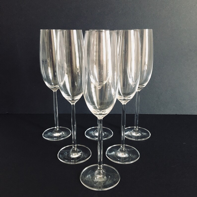 May include: Six clear glass champagne flutes arranged in a semi-circle on a black surface.