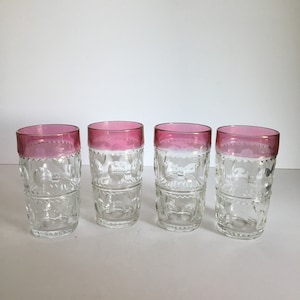 May include: Four clear drinking glasses with a pink rim and decorative etched pattern. The glasses are tall and cylindrical, suitable for water or other beverages. The pink rim adds a pop of color to the glassware.