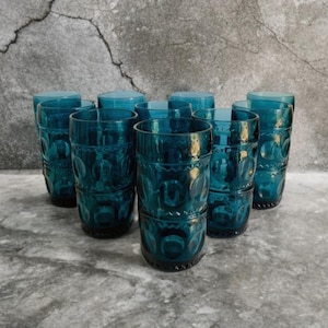 May include: A set of eight teal-colored drinking glasses. Each glass has a cylindrical shape with a decorative pattern of circles and lines. The glasses are arranged in a group, showcasing their design and color.