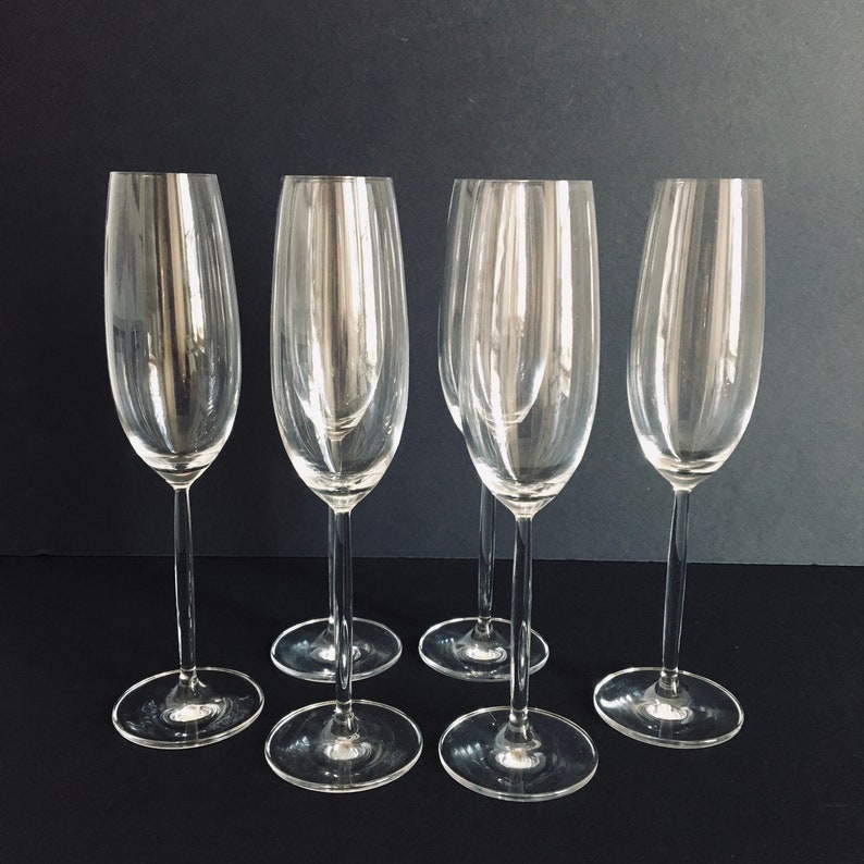 May include: Five clear glass champagne flutes with a tall, slender design. The flutes are arranged in a semi-circle on a black surface.