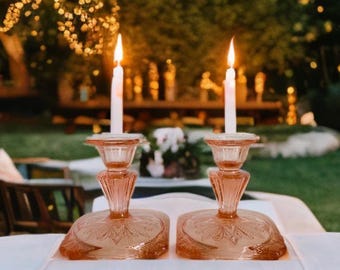 May include: Two lit candles sit atop matching pink glass candle holders with ornate bases. The candle holders are on a white tablecloth, with a blurred outdoor background of trees and lights.