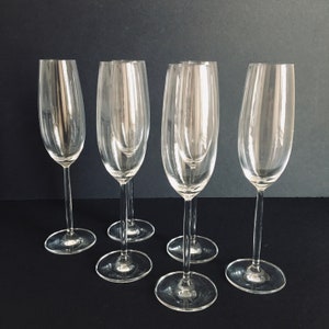 May include: Five clear glass champagne flutes with a tall, slender stem and a bowl that is slightly wider at the top. The flutes are arranged in a row on a black surface.