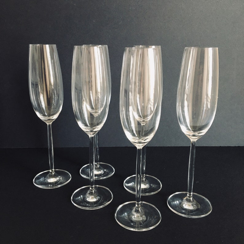May include: Five clear glass champagne flutes with a tall, slender stem and a bowl that is slightly wider at the top. The flutes are arranged in a row on a black surface.