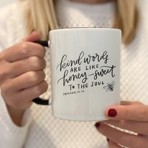 May include: White ceramic mug with a black handle. The mug has a black handwritten quote that reads "Kind words are like honey-sweet to the soul" with the scripture reference "Proverbs 16:24" below. A small black bee illustration is to the right of the quote.
