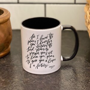 May include: White ceramic mug with a black rim and handle. The mug has black lettering that reads "for I know the plans I have for you, declares the Lord, "plans to prosper you not to harm you, plans to give you a hope & a future." Jeremiah 29:11