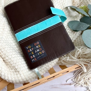 May include: A brown leather journal with a turquoise blue ribbon closure. A sticker with the text "YOU GO BEFORE ME AND BEHIND ME" and the scripture reference "Joshua 1:9" is on the front cover.