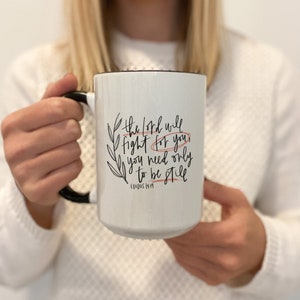 Op de afbeelding: Een witte keramische mok met een zwarte handgreep en een zwarte rand. De mok heeft een zwart-wit ontwerp met de tekst "the Lord will fight for you, you need only to be still, EXODUS 14:14".