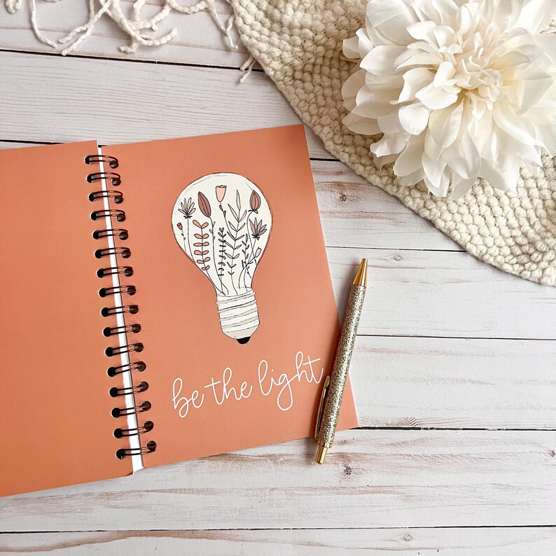 Puede incluir: Un cuaderno de espiral de color melocot&oacute;n con una ilustraci&oacute;n de bombilla llena de flores y el texto "be the light" en escritura blanca.