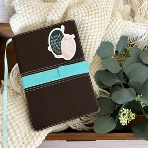 May include: A brown leather-bound journal with a turquoise blue ribbon bookmark. A sticker with the text "Be strong and courageous. Do not be afraid; do not be discouraged, for the Lord your God will be with you wherever you go." is attached to the front cover.