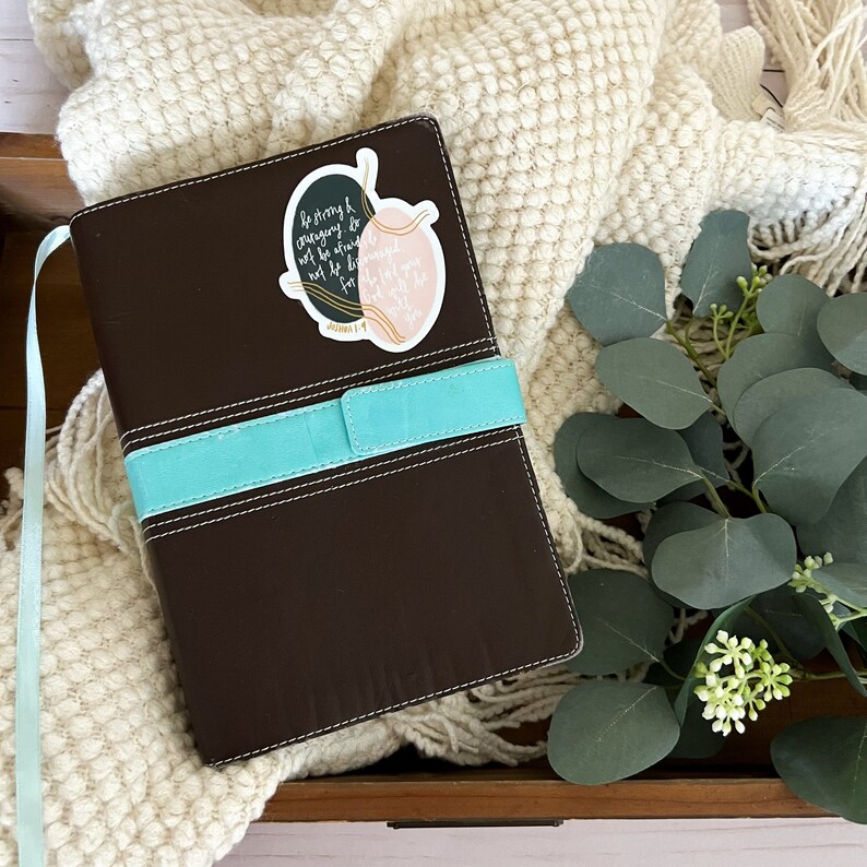May include: A brown leather-bound journal with a turquoise blue ribbon bookmark. A sticker with the text "Be strong and courageous. Do not be afraid; do not be discouraged, for the Lord your God will be with you wherever you go." is attached to the front cover.