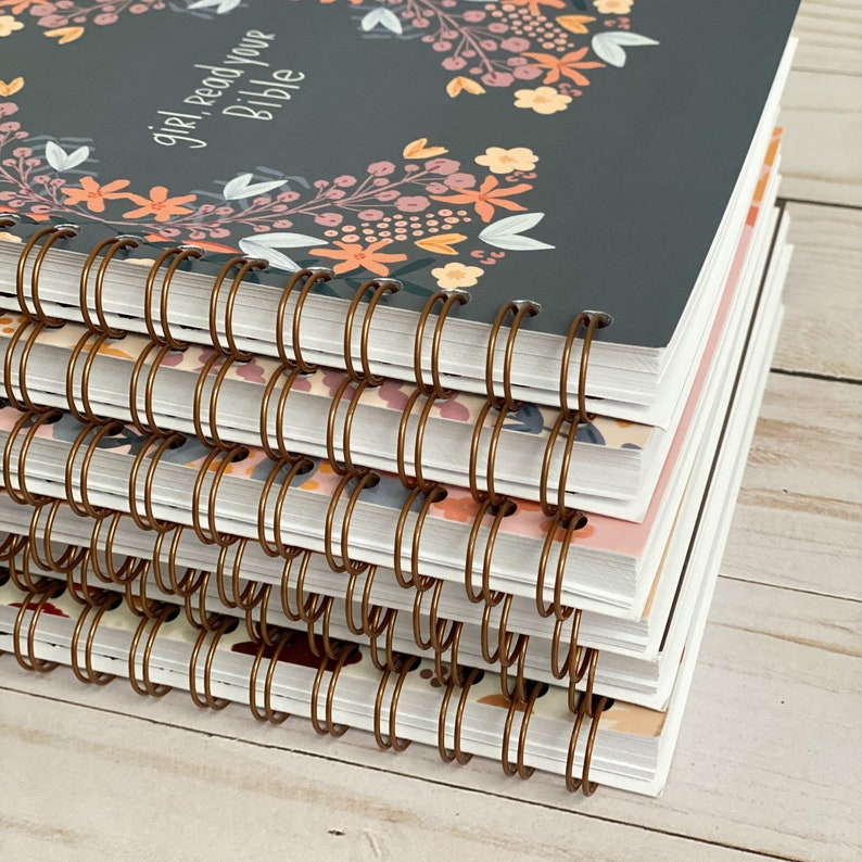 May include: A stack of spiral bound notebooks with a floral design and the text "girl, read your Bible" on the top notebook. The notebooks have copper colored binding.