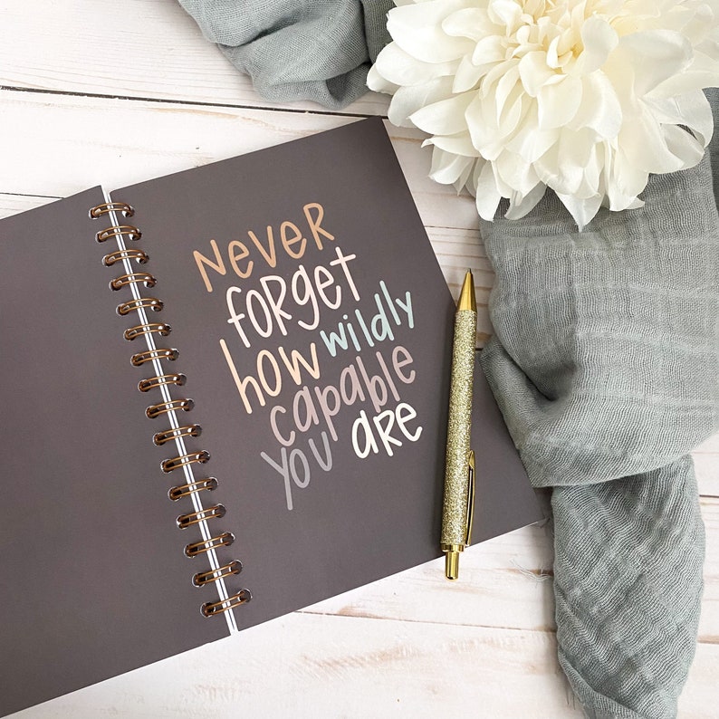 May include: A grey spiral bound journal with the words "Never forget how wildly capable you are" written in colourful script. A gold pen is lying on top of the journal.