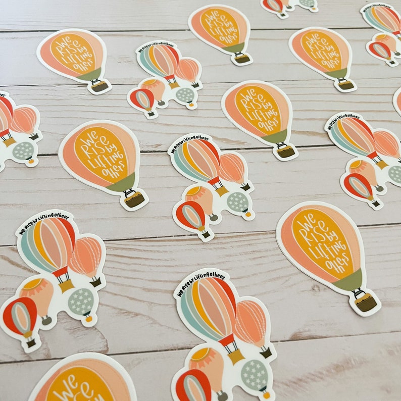May include: A set of colourful hot air balloon stickers with the text "We Rise by Lifting Others". The stickers are on a light wood background.