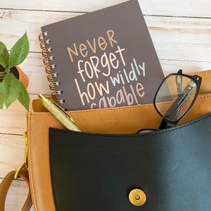 May include: A brown leather bag with a black flap and a gold snap closure. Inside the bag is a grey spiral notebook with the text "Never forget how wildly capable" written in pink, white, and brown. A gold pen and a pair of black reading glasses are also inside the bag.
