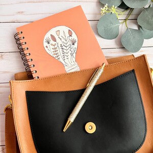 Puede incluir: Una bolsa de cuero marr&oacute;n claro con un bolsillo negro y un bol&iacute;grafo dorado en su interior. La bolsa tiene un cierre de bot&oacute;n dorado. Una libreta rosa con encuadernaci&oacute;n en espiral est&aacute; en el bolsillo. La libreta tiene un dibujo de una bombilla con flores dentro en la portada.