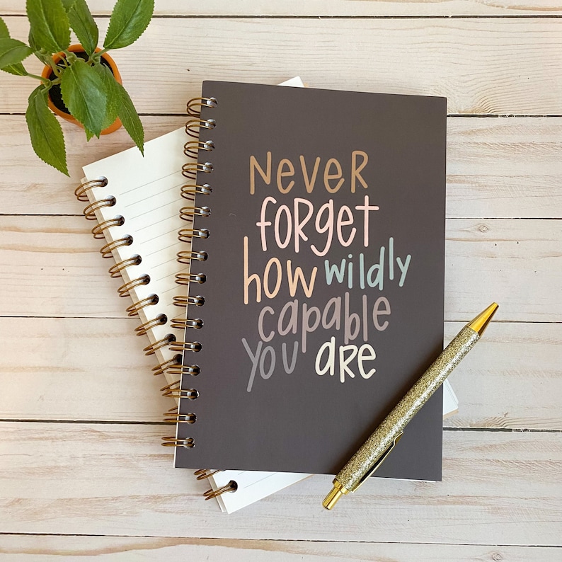 May include: A grey spiral bound journal with the text "Never forget how wildly capable you are" in colourful lettering. The journal is on a wooden surface with a gold pen.