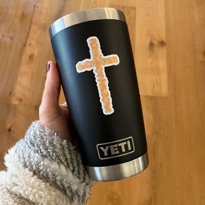 May include: A black Yeti tumbler with a floral cross sticker and the word "YETI" printed on the side.