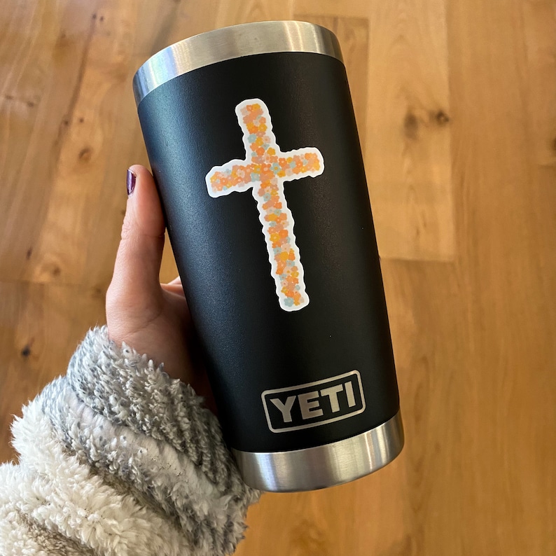 May include: A black Yeti tumbler with a floral cross sticker and the word "YETI" printed on the side.